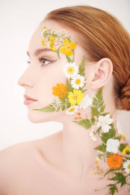 Çekici kızıl saçlı, zarif makyajlı ve yüzünde ve omzunda yaz çiçekleri olan bir kızın yakın çekimi. Beyaz arka plan. Kozmetik ve parfüm. Yazın kokusu..