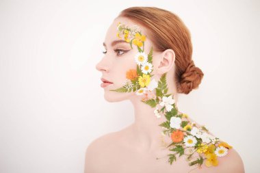 Yazın kokusu. Zarif makyajlı, yüzünde ve omzunda yaz çiçekleri olan çekici kızıl saçlı bir kız beyaz arka planda poz veriyor. Kozmetik ve parfüm. 