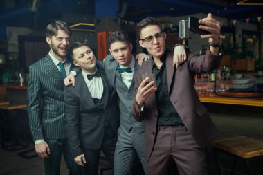 Klasik takım elbiseli yakışıklı genç erkekler bir barda rahatlar ve grup selfie 'si çekerler. Erkek modası. İş adamları kulübü.