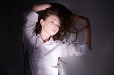 Beyaz gömlekli güzel bir kız, uzun kahverengi saçlarıyla gölgelerin ve ışıkların arasında koyu bir arka planda oynuyor..