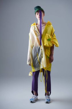 Parlak sarı ceketli, mor eşofmanlı ve parlak mor saçlı yeşil şapkalı bir erkek mankenin moda fotoğrafı. Gençlik tarzı ve yaratıcı tasarımcı kıyafetleri. Tam boy portre.