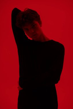 Ergenler ve iç dünya. Dalgalı siyah saçlı, yüzünde depresif bir ifadeyle ayakta duran ve kendine sarılan yakışıklı bir adamın portresi. Kırmızı arkaplanda stüdyo çekimi. 