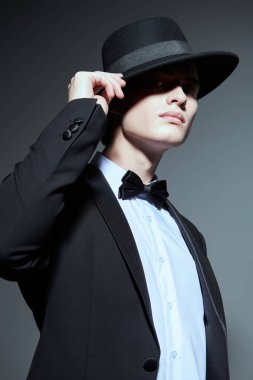 Siyah smokinli yakışıklı bir genç. Papyonlu ve şapkalı. Gri stüdyo arka planında gölgesi var. Erkek modası ve güzelliği. 
