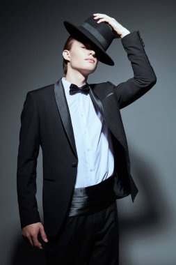 Gri bir stüdyo arka planında poz veren şık siyah smokinli, papyonlu ve şapkalı genç yakışıklı bir adamın portresi. Erkek modası ve güzelliği. 