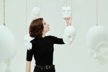 Siyah elbiseli bir kız, beyaz bir odada farklı maskelerle ayakta duran farklı maskeler deniyor. İnsan rolleri. İkiyüzlülük. Zihinsel bozukluklar.