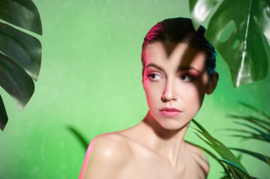 Yumuşak makyajlı hoş bir kızın portresi tropik yeşilliklerin arasında duruyor. Yeşil arka plan ve pembe ışık. Orman fotoshop 'u. Spa, cilt bakımı ve kozmetik ürünleri. 