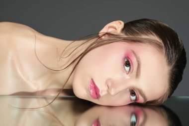 Açık pembe makyajlı güzel model kadın yansıtıcı yüzeyde uzanan kameraya bakıyor. Stüdyo çekimi. Kadın güzelliği ve sağlığı. Kozmetik ve cilt bakımı. 