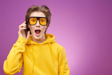 Şık güneş gözlüklü ve sarı kapüşonlu genç bir çocuk telefondan duyulan habere duygusal tepki veriyor. Gençlik yaşam tarzı, iletişim. Modern gençler işte. Kopyalama alanı olan mor arkaplan.