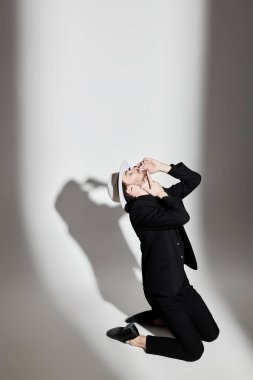 Şık siyah takım elbiseli zarif bir esmer adam ve beyaz şapkalı beyaz bir adam ışık huzmesi tarafından aydınlatılan beyaz bir duvara karşı dizlerinin üzerinde duruyor. Moda fotoğrafı. Işık ve gölge.