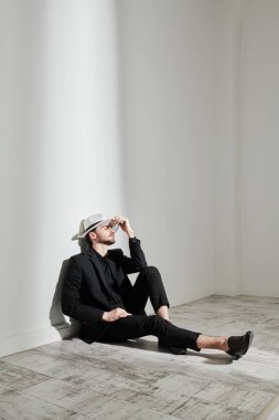 Erkek modası. Siyah takım elbiseli zarif giyinmiş esmer bir adam ve beyaz şapkalı, gözleri kapalı, beyaz bir duvara yaslanmış bir şekilde oturuyor. Tam boy stüdyo resmi..