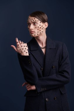 Yüzünde ve elinde metal çivilerle poz veren koyu mavi ceketli çekici bir gencin stüdyo fotoğrafı. Koyu mavi arka plan. Moda fotoğrafı.. 