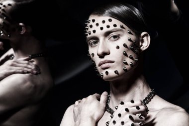 Yüzünde ve elinde metal çiviler olan yakışıklı bir esmer adam aynadaki yansımasının arka planında poz veriyor. Moda ve sanat. 