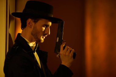 Siyah paltolu cesur bir esmer adam portresi ve elinde bir silahla, bir ışık huzmesi ile karanlık odada duran bir şapka. Retro noir tarzı. Suç dünyası, mafya..