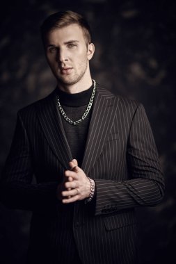 Siyah kazaklı ve ceketli yakışıklı, ciddi sarışın bir adam koyu bir arka planda duruyor. Erkek modası.