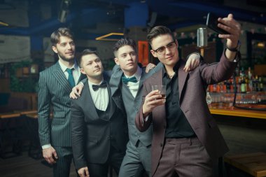 Klasik takım elbiseli yakışıklı genç erkekler bir barda rahatlar ve grup selfie 'si çekerler. Erkek modası. İş adamları kulübü.