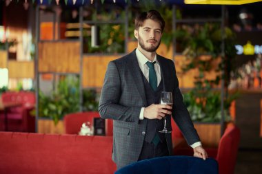 Zarif klasik takım elbiseli, kendine güvenen yakışıklı bir esmer bir adam elinde bir bardak şarapla bir restoranda duruyor. Erkek güzelliği, moda. İş stili. İşadamı. 