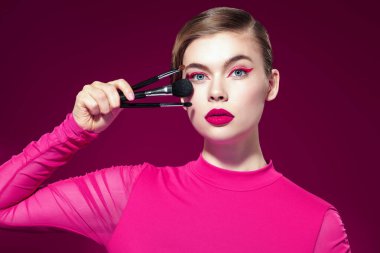 Parlak pembe rujlu, pembe okları olan, fuşya arka planında çeşitli makyaj fırçaları tutan güzel bir sarışın kız. Makyaj ve makyaj malzemeleri. Makyaj araçları.
