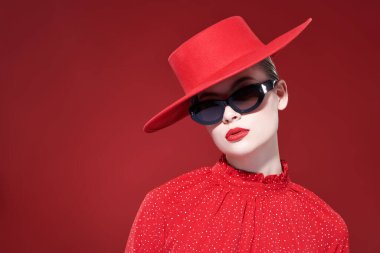 Moda ve güzellik. Parlak makyajlı, şık, kırmızı elbiseli, kırmızı şapkalı ve şık güneş gözlüklü güzel bir manken. Kopyalama alanı olan kırmızı arkaplan. 