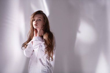 Beyaz gömlekli, uzun kahverengi saçlı güzel bir kız gölgeli beyaz bir arka planda sakince ve dalgın bir şekilde duruyor. Boşluğu kopyala.