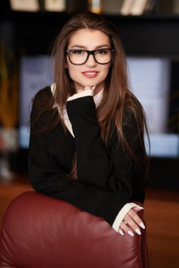 Çarpıcı, kendine güvenen bir iş kadını modern bir ofiste deri koltuğun yanında duruyor. Başarılı genç insanlar. Modern iç mekan, mobilya.