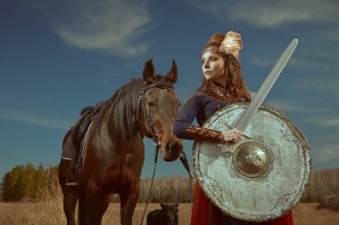 Cesur ve güzel kadın savaşçı Valkyrie savaş alanında savaş atının yanında elinde bir kılıç ve kalkanla duruyor. İskandinav mitolojisi. Efsanevi fantezi.