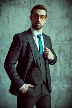 Moda fotoğrafı. Üç parçalı şık takım elbiseli zarif yakışıklı bir adamın portresi ve gri bir grunge duvarın arkasında poz veren bir kravat.. 