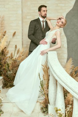Yakışıklı damat zarif beyaz elbisesiyle sevgili gelinine sarılıyor. Tam boy portre. Düğün modası.