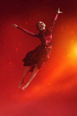 Dans sanatı, modern bale. Kırmızı kadife elbiseli güzel balerin kız stüdyodaki kırmızı arka planda atlıyor. Tam boy portre.