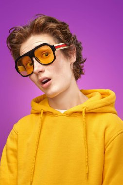 Parlak sarı kapüşonlu duygusal bir genç kameraya şaşkınlıkla bakıyor. Parlak mor arka plan. Eğitim. Gençlik hayatı, başarı. Duygular.