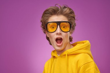 Parlak genç kıyafetleri ve şık güneş gözlükleriyle kameraya büyük bir sürprizle bakan duygusal bir genç. Mor arka plan. Gençlik yaşam tarzı ve duyguları. 