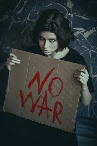 stock image Anti-war protest. Portrait of a brunette girl in black clothes sitting by a grunge wall with the sign 