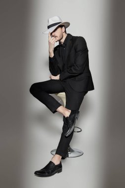 Zarif bir esmer adam portresi şık siyah bir takım elbise ve beyaz bir şapka içinde beyaz bir duvarın kenarında ışık huzmesi ile aydınlatılmış bir taburede oturuyor. Moda fotoğrafı. Işık ve gölge.