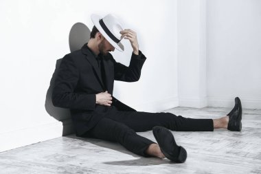 Erkek modası. Siyah takım elbiseli ve beyaz şapkalı zarif giyinmiş esmer bir adam beyaz bir duvara yaslanmış yerde oturuyor. Tam boy stüdyo resmi..