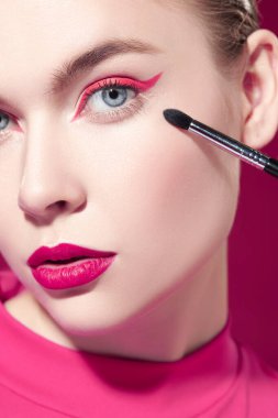 Parlak pembe rujlu ve gözlerinde pembe oklar olan güzel bir model kız fuşya arka planında fırçayla makyaj yapıyor. Güzellik, makyaj ve kozmetik. Makyaj araçları.