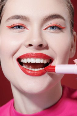 Sevimli kırmızı renk. Güzel, gülümseyen bir kız modelin parlak kırmızı ruj sürerken çekilmiş bir fotoğrafı. Yakın çekim. Makyaj ve kozmetik. 