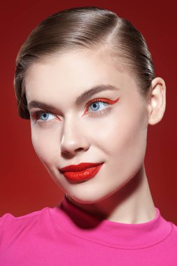 Güzellik konsepti. Kırmızı arka planda kırmızı okları ve parlak kırmızı rujuyla gülümseyen güzel bir sarışın kız poz veriyor. Makyaj ve kozmetik. 