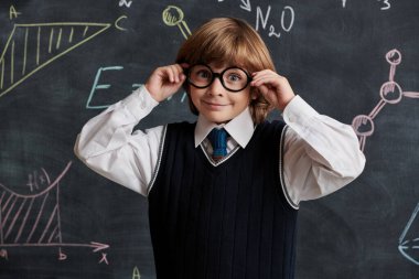 Güzel bir okul üniforması ve gözlüklü yakışıklı bir çocuk kimyasal formüllerle tahtada duruyor ve mutlu bir şekilde gülümsüyor. Eğitim. Akıllı çocuklar..