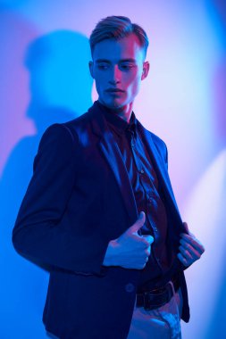 Şık gömlek, ceket ve pantolon giymiş çekici, genç bir sarışının moda portresi. Duvarın yanında çok renkli bir ışıkla duruyor. Erkek modası.