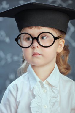 Beyaz gömlekli, akademik şapkalı ve gözlüklü zeki bir kız formüller ve diyagramlarla arka planda duruyor. Çocuklar ve eğitim. 