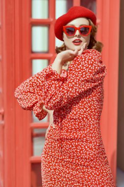 Güzel kırmızı elbiseli, kırmızı bereli ve zarif kırmızı gözlüklü çekici bir kız bir telefon kulübesinin önündeki caddede duruyor. Güzellik, moda. Retro pin-up biçimi.