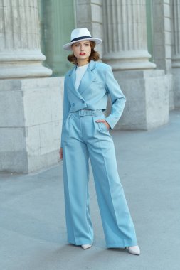 Hollywood tarzı. Güzel kahverengi saçlı bir kadının portresi zarif bir gökyüzü mavi takım elbise ve beyaz şapkalı, bir şehir caddesinde duruyor. 40 'lı yılların tarzı. Makyaj ve saç stili.