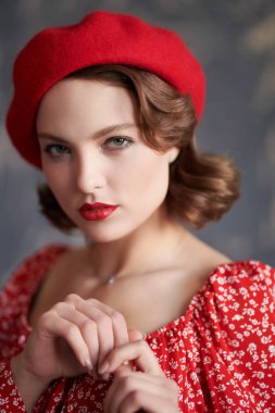 Kadınlık ve retro stil. Zarif makyajı ve saç stili olan güzel bir genç bayan stüdyoda kırmızı elbise ve kırmızı bereyle poz veriyor. Güzellik, moda.