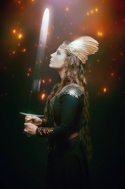 Kanatlı bir kaskla elinde kılıçla duran güzel bir kadın Valkyrie 'nin yan görüntüsü. Stüdyo portresi karanlık bir arka planda, ateş kıvılcımları var. İskandinav mitolojisi. Efsanevi fantezi.