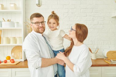 Mutlu aile fotoğrafı. Mutlu anne ve baba sıcacık bir mutfakta durup kızlarını ellerinde tutuyorlar ve gülümsüyorlar. Modern iç mekan, mutfak mobilyası.. 