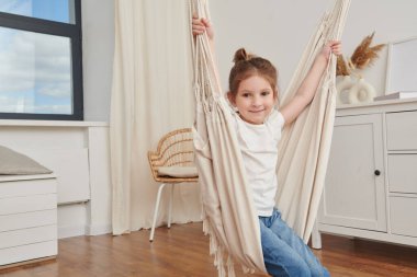 Sevimli bir kız çocuğu, rahat bir yatak odasında salıncakta neşeyle sallanır. Çocuk odası tasarımı. Mutlu çocukluk.