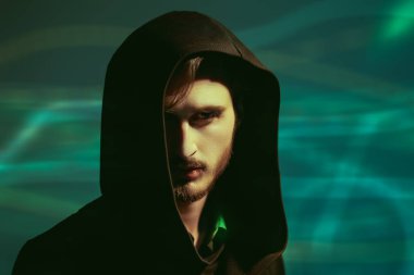 Siyah bir başlığın altından kameraya dikkatle ve dikkatle bakan esmer bir adamın stüdyo portresi. Yeşil arka plan. İnsanlar ve duygular. 