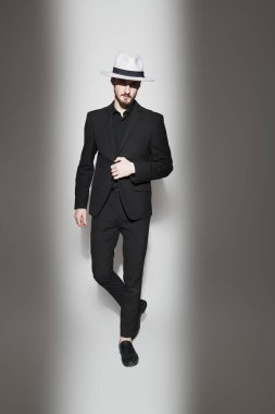 Zarif siyah takım elbiseli iyi giyimli esmer bir adam ve parlak bir ışıkta beyaz bir arka planda duran bir şapka. Tam boy stüdyo resmi. Zarif erkek modası. Işık ve gölge.