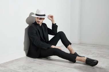 Siyah takım elbiseli, beyaz şapkalı şık bir genç beyaz bir duvara yaslanmış yerde oturuyor. Tam boy stüdyo resmi. Erkek modası. 