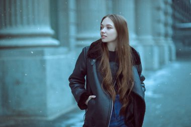 Deri ceketli ve kot pantolonlu güzel modern bir kız karlı bir kış gününde şehir caddesinde yürüyor. Gençler, yaşam tarzı..