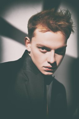 Siyah smokinli sarışın genç bir adam açık bir arka planda poz veriyor. Kameraya şüpheyle ve güvensizlikle bakıyor. Erkek modası ve güzellik.